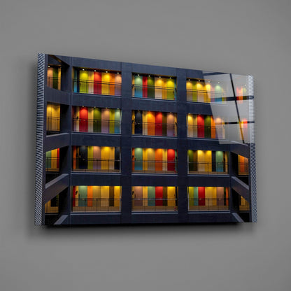 Modern glass wall art featuring a multi-story building facade with colorful illuminated window panels in red, yellow, green, and orange against dark framework.