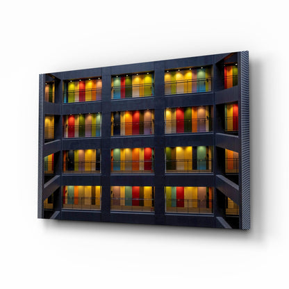 Modern dark facade with four stories of illuminated balconies featuring vibrant multicolored glass panels in warm and cool tones.