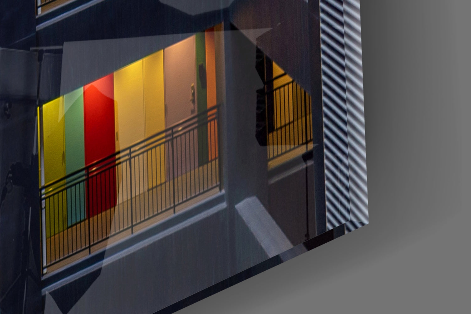 Colorful glass wall art with yellow, green, red, and orange panels mounted on a modern building facade with metal railings and shadow effects.