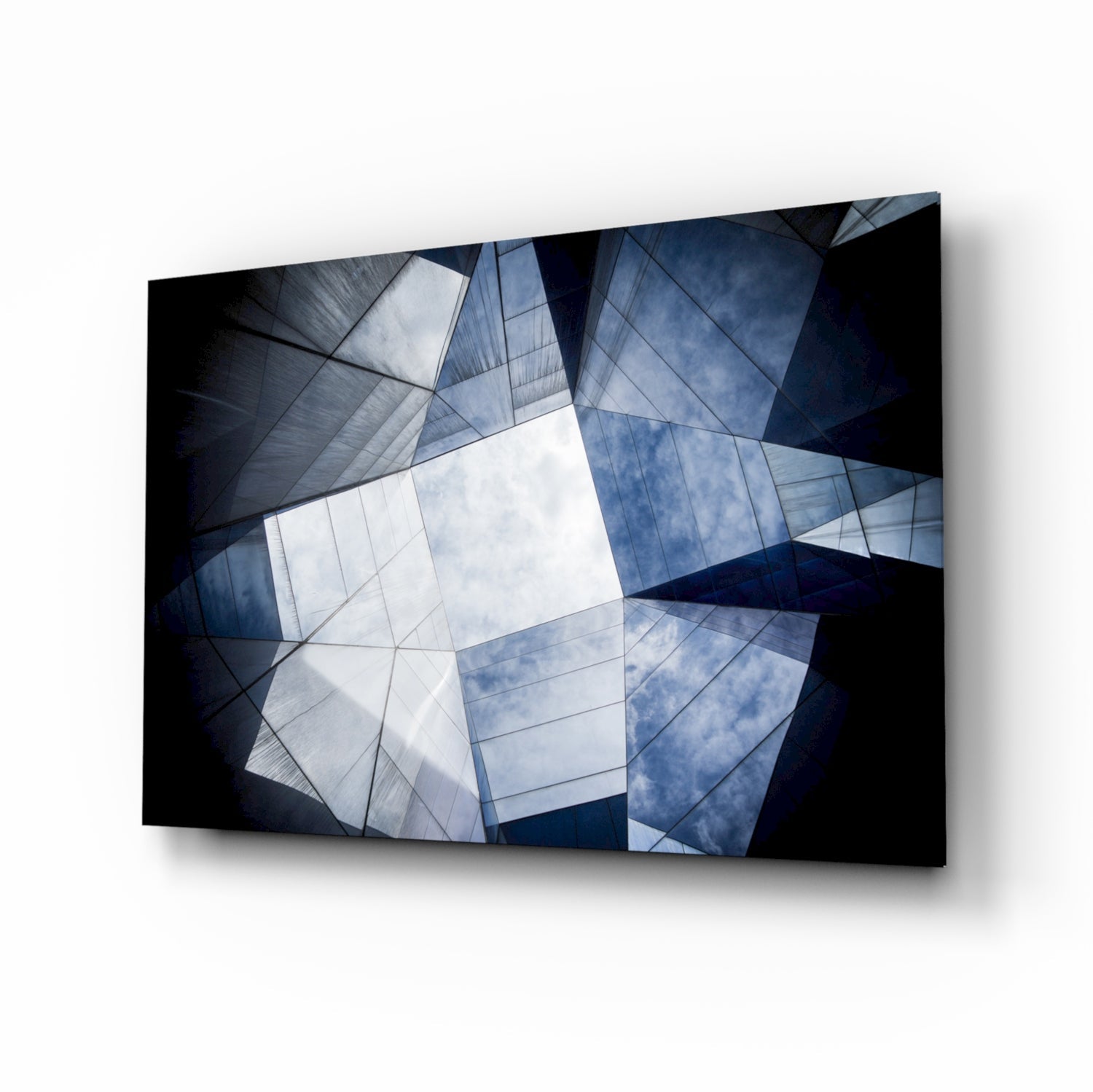Abstract geometric glass wall art displaying overlapping angular panels in blue, white, and black tones with sky reflections, mounted at an angle against white background.