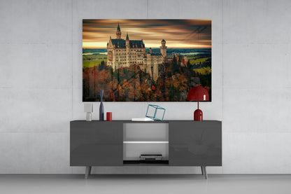 Castle glass wall art displaying Neuschwanstein Castle at sunset, mounted above gray media console with red decorative accessories.