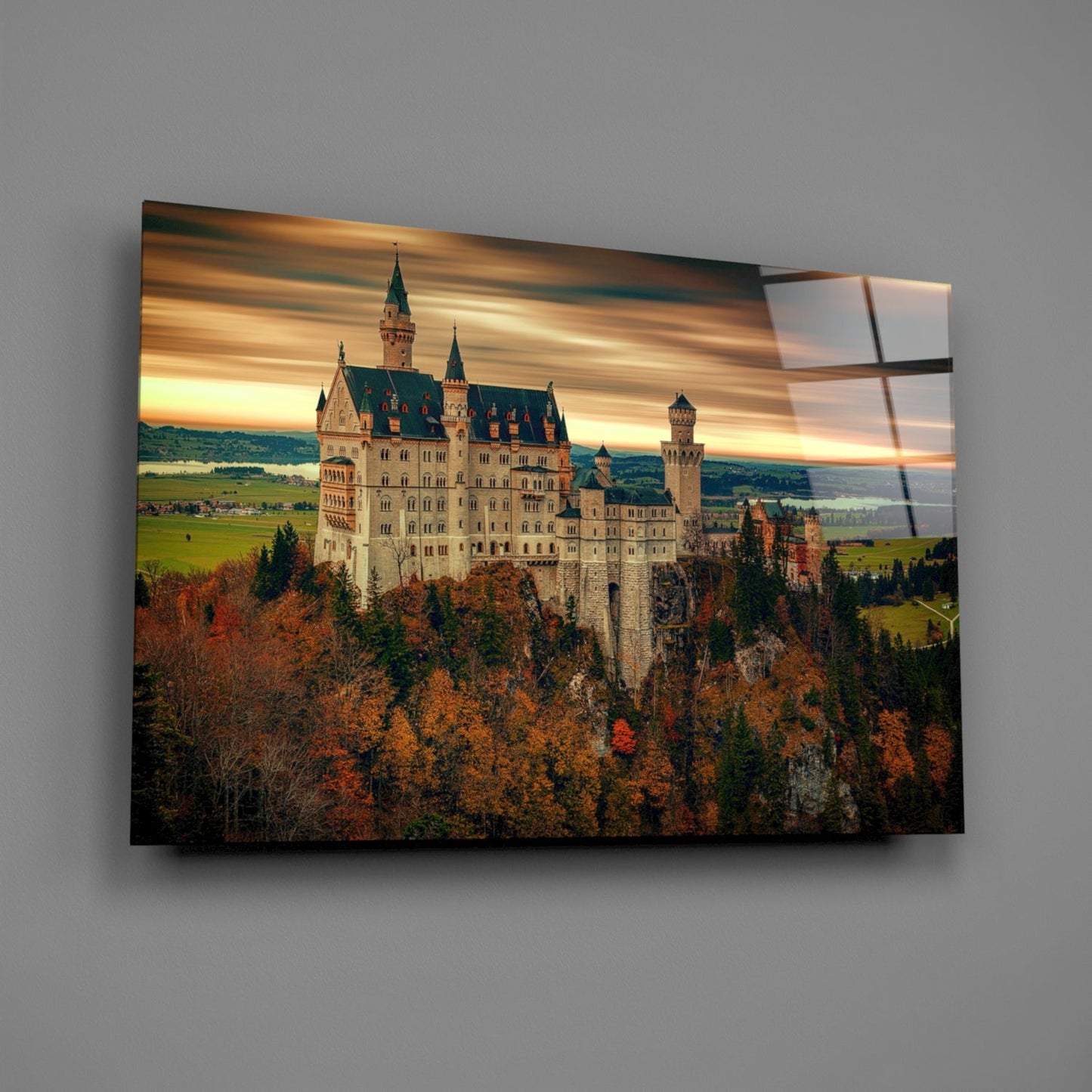Neuschwanstein Castle on glass art panel mounted on gray wall, showing autumn forest landscape with golden sunset sky.