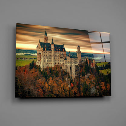 Neuschwanstein Castle on glass art panel mounted on gray wall, showing autumn forest landscape with golden sunset sky.