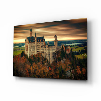 Neuschwanstein Castle perched on a forested mountainside at sunset, displayed as a glass wall art panel with autumn foliage below.