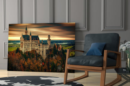 Castle Glass Wall Art displaying Neuschwanstein Castle at sunset, mounted on a light grey paneled wall beside a wooden chair with blue cushion.