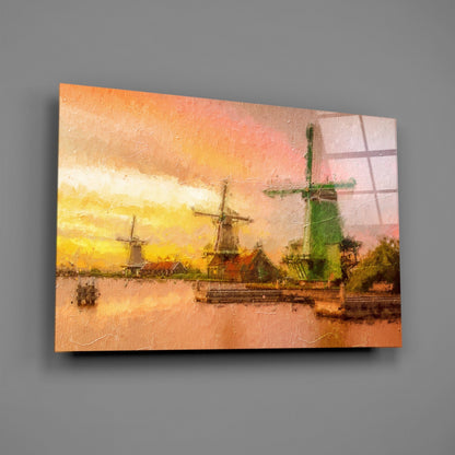 Glass wall art depicting Dutch windmills by a waterway at sunset in warm orange and yellow tones, displayed at an angle against gray background.