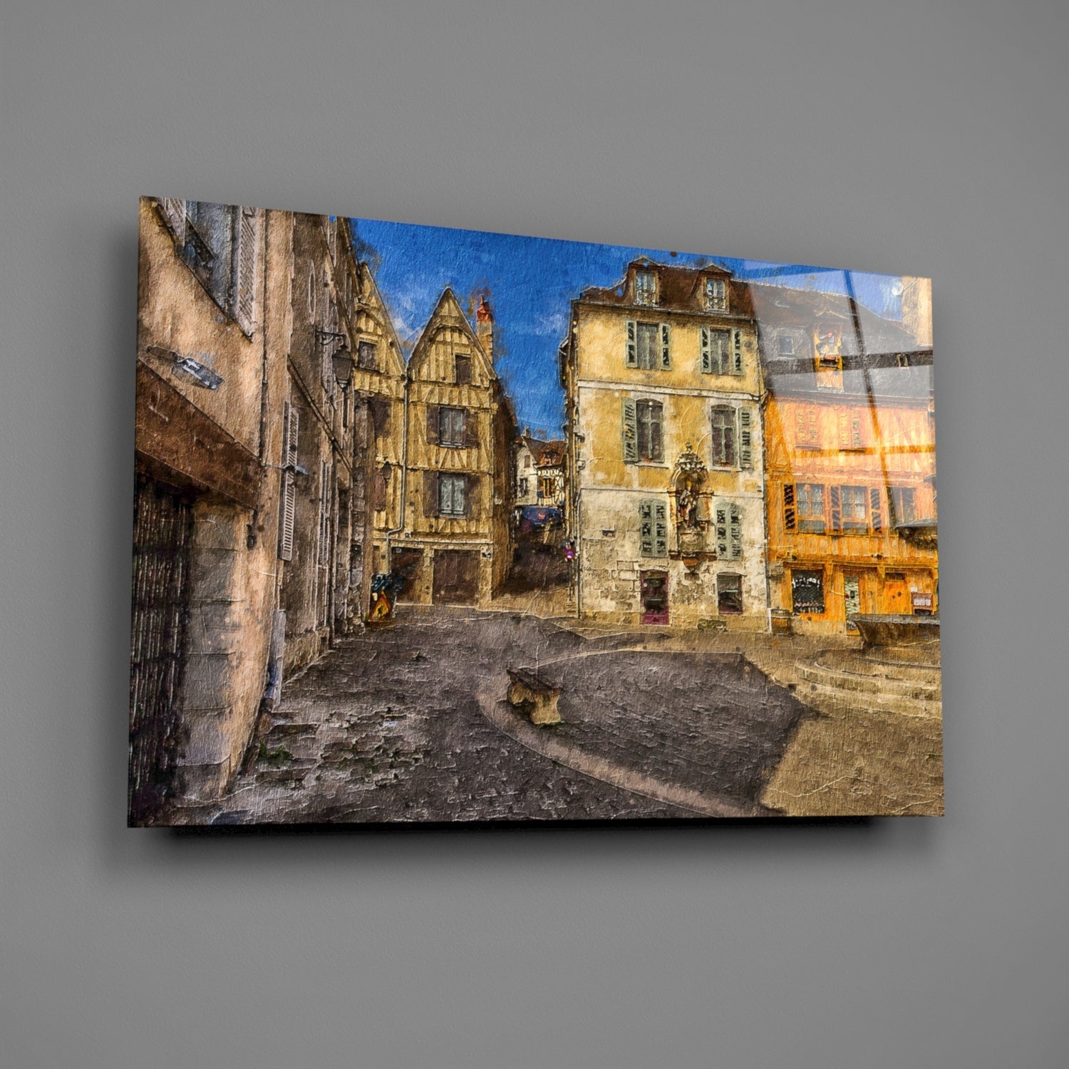 Glass wall art print displayed at an angle showing a medieval European square with half-timbered buildings in yellow and cream tones under blue sky.