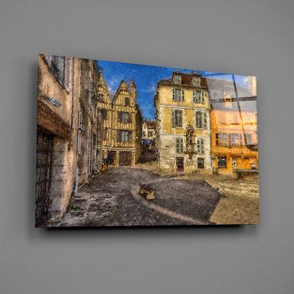 Glass wall art print displayed at an angle showing a medieval European square with half-timbered buildings in yellow and cream tones under blue sky.