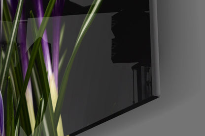 Purple flowers glass wall art displayed on gray wall with green plant foliage in foreground.