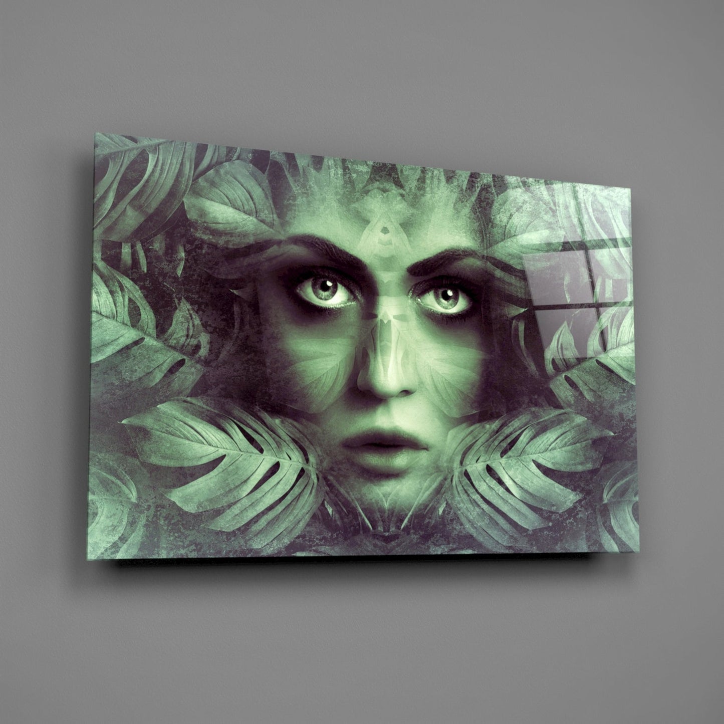 Glass wall art featuring a woman's face with striking eyes surrounded by green tropical leaves in monochromatic tones, displayed at an angle against gray background.