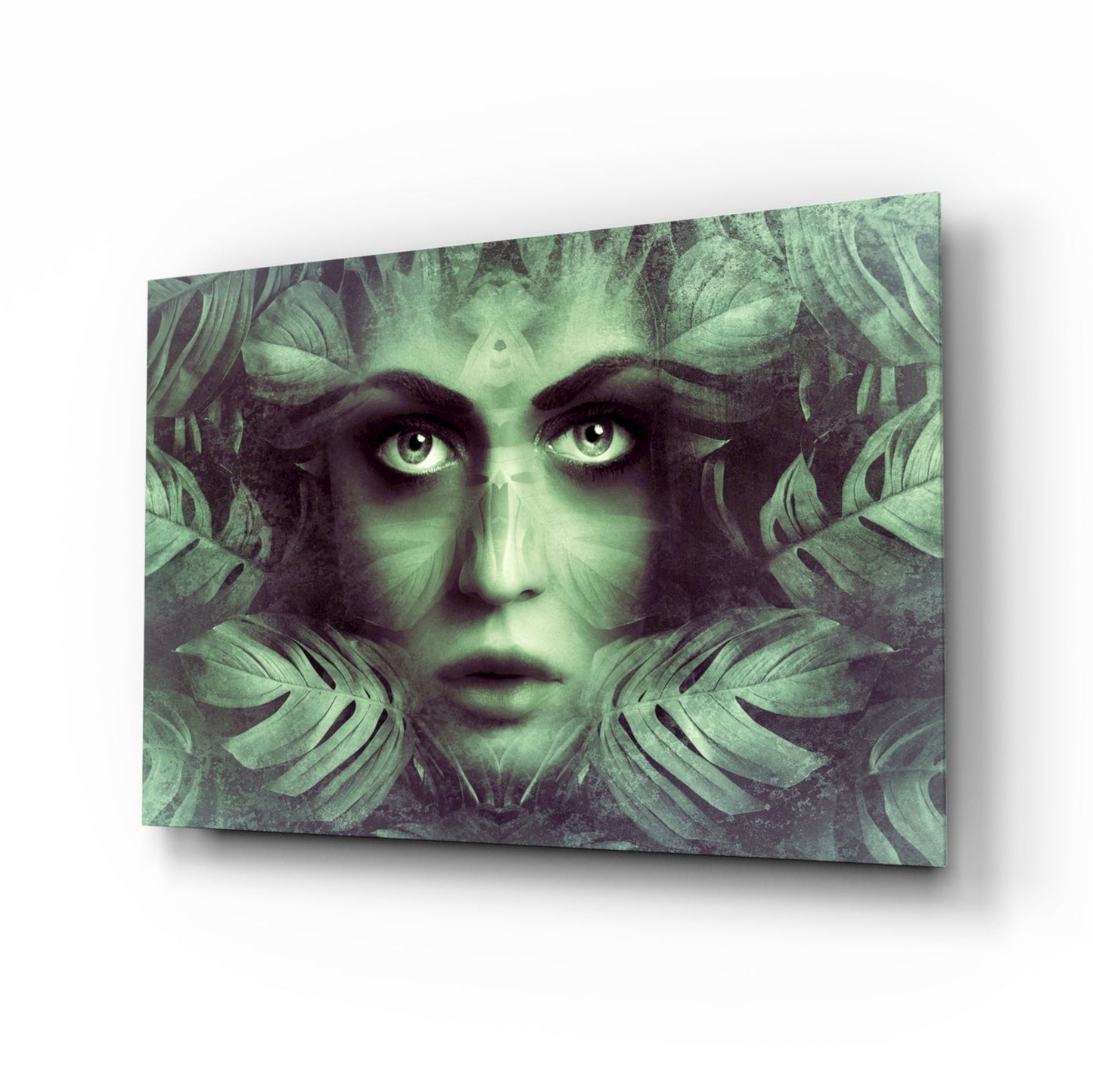 Angled view of glass wall art featuring a woman's face with pale skin and wide eyes surrounded by green tropical leaves in monochromatic tones.