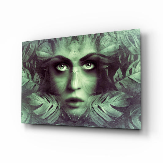 Angled view of glass wall art featuring a woman's face with pale skin and wide eyes surrounded by green tropical leaves in monochromatic tones.