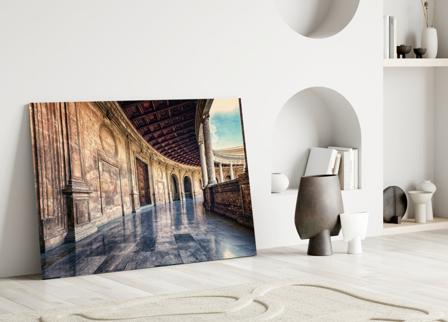 Glass wall art depicting a sunlit baroque colonnade with ornate architecture and blue reflective floor, leaning against white wall beside decorative vessels.