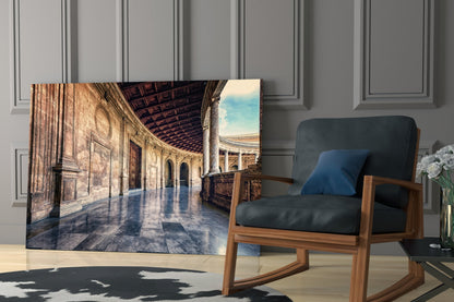 Glass wall art displaying a sunlit baroque colonnade with ornate architecture and reflective marble floors, displayed in a modern living room next to a wooden chair.