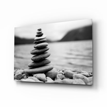 Stacked river stones balanced in a pyramid on a pebbly beach with blurred water and mountains in the background, displayed as a framed black and white glass wall art print.