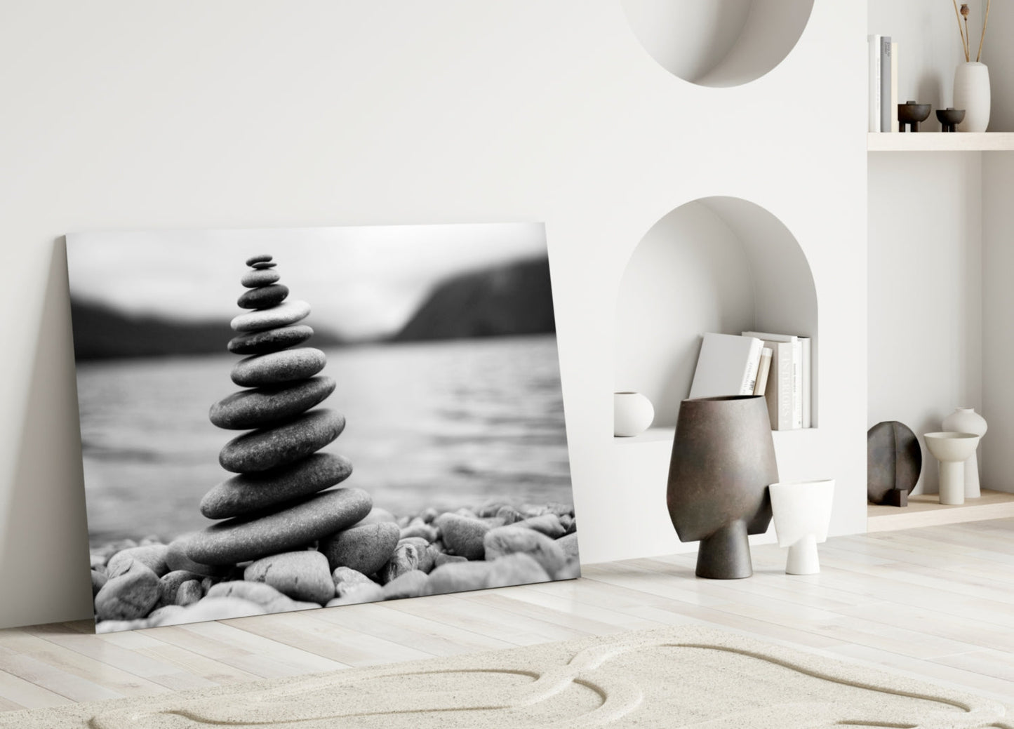 Black and white glass wall art depicting a stacked stone cairn on a beach, leaning against a white wall in a minimalist interior setting.