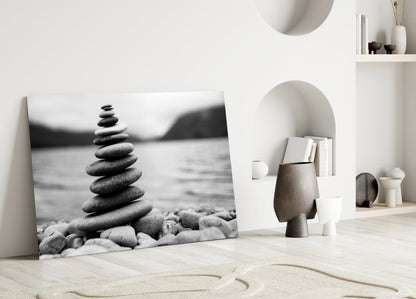 Black and white glass wall art depicting a stacked stone cairn on a beach, leaning against a white wall in a minimalist interior setting.