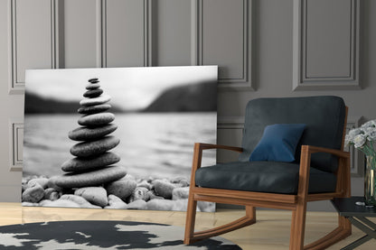 Black and white glass wall art depicting stacked stones by water, displayed in a modern living room with wooden chair and blue pillow.