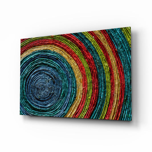 Angled perspective view of multicolored concentric woven rings spiral glass wall art with blue, teal, red, green, and gold thread details.