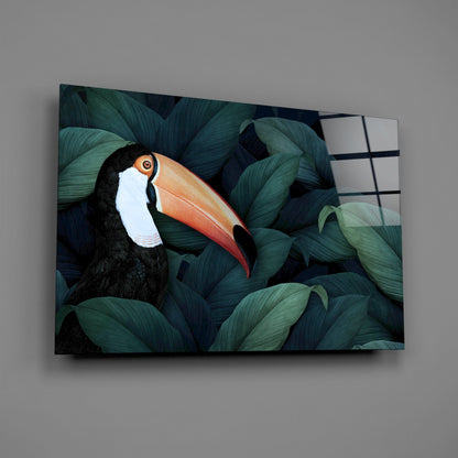 Toucan with orange beak and white face nestled among dark teal leaves on glass wall art, displayed at angle against gray wall.
