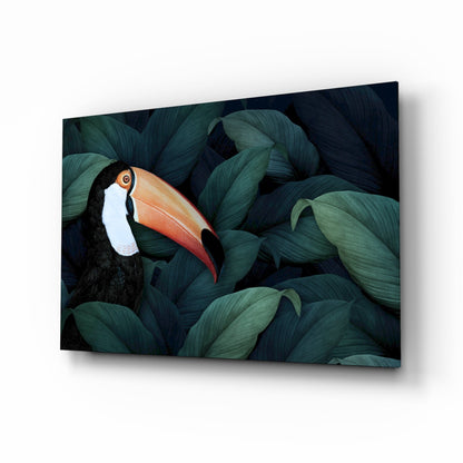 Angled view of glass wall art featuring a toucan with black plumage and orange beak nestled among dark green foliage.