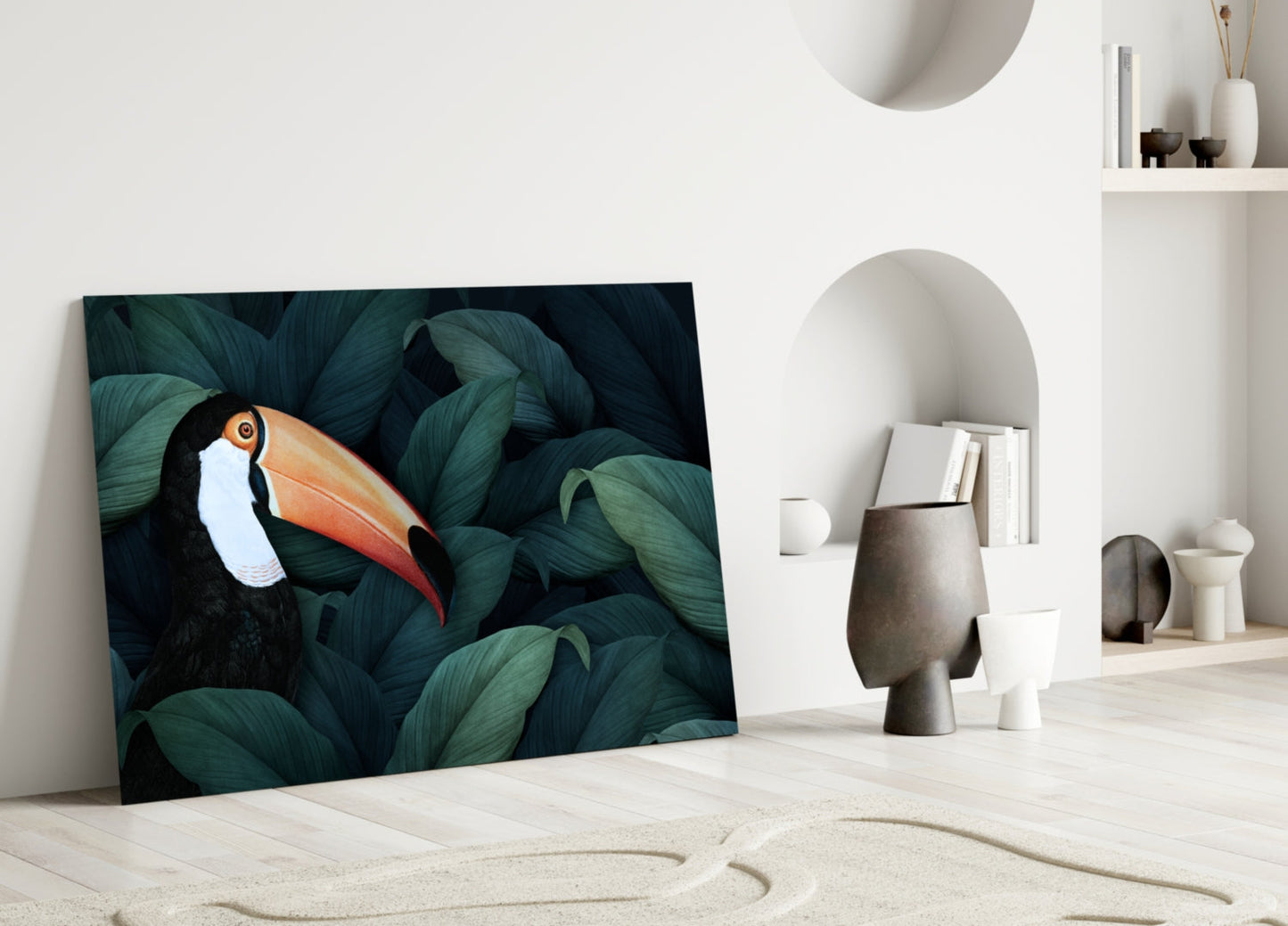 Toucan glass wall art leaning against white wall in minimalist interior with decorative objects and shelving.