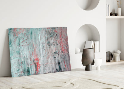 Abstract glass wall art with turquoise, pink, and gray brushstroke pattern leaning against white wall in minimalist interior.