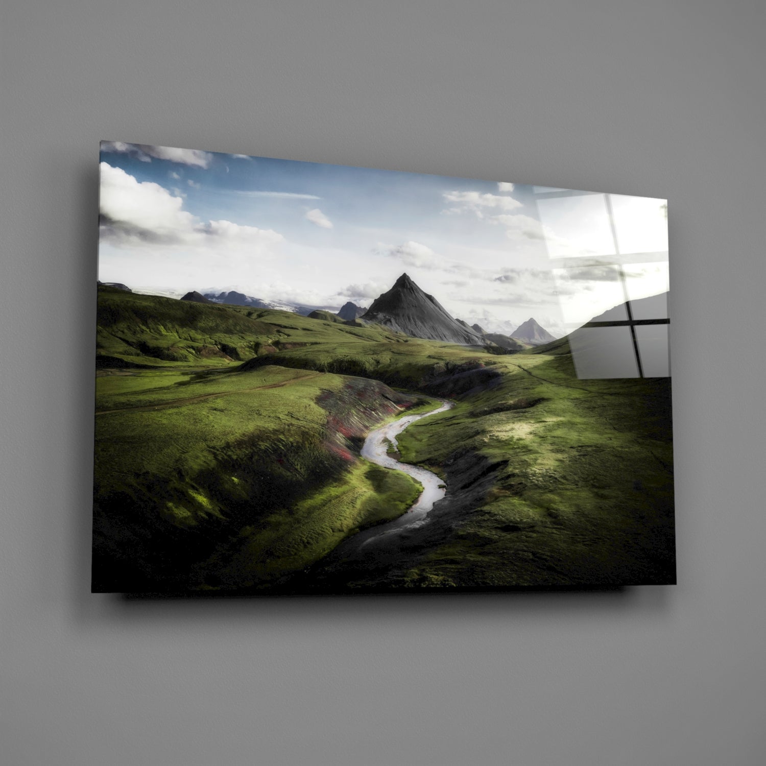 Angled view of glass wall art displaying lush green valley with winding river and mountain peak under cloudy sky.