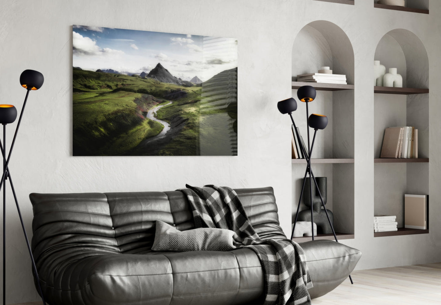 Glass wall art depicting a green valley landscape with mountains, river, and blue sky, displayed above a black leather sofa in a modern living room.
