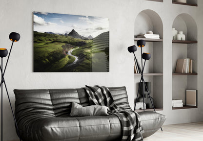 Glass wall art depicting a green valley landscape with mountains, river, and blue sky, displayed above a black leather sofa in a modern living room.