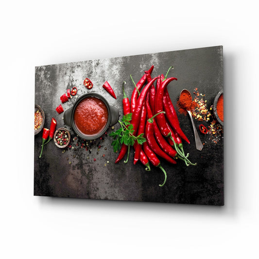 Glass wall art panel displaying fresh red chili peppers, spices, and sauce arranged on a dark textured surface, shown at an angled perspective.