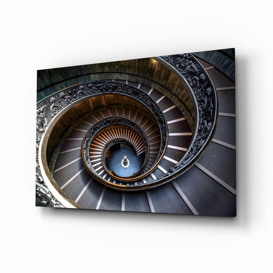 Aerial view of a spiral staircase with ornate carved railings, descending through concentric circles in brown, gold, and gray tones.