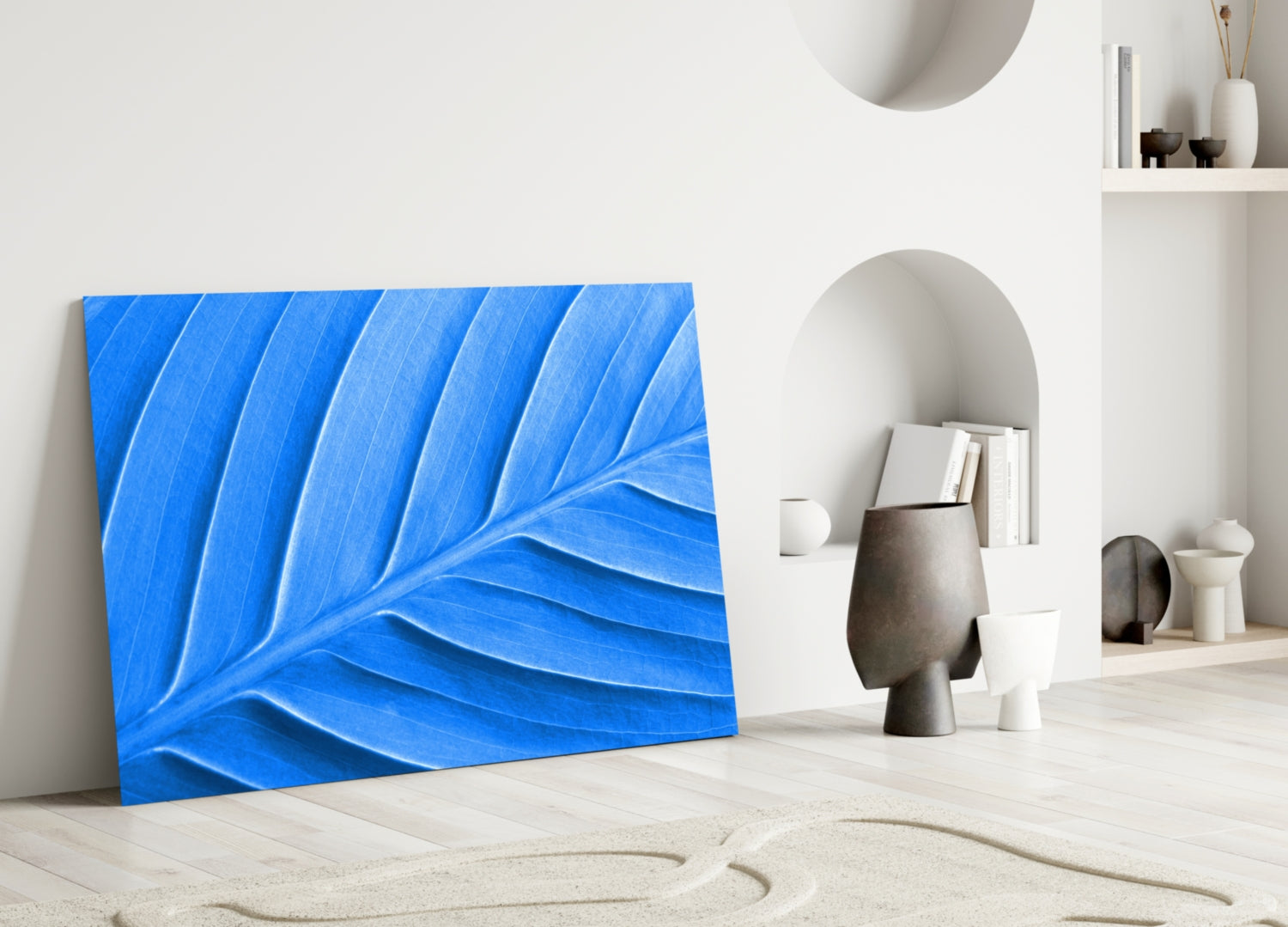 Blue leaf-patterned glass wall art leaning against white wall in minimalist interior setting with decorative objects.