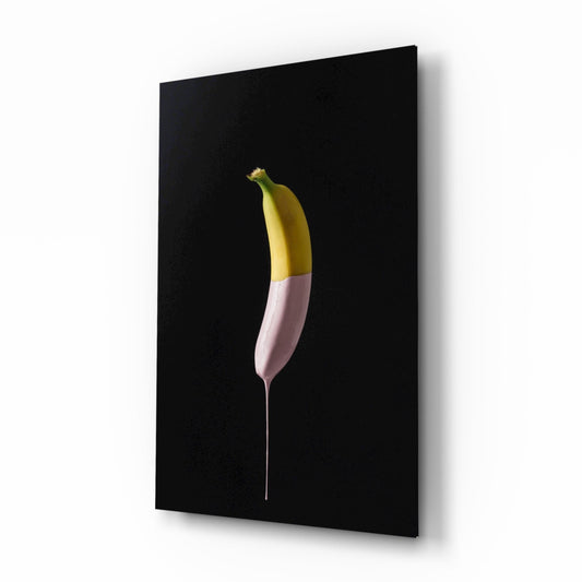 Angled view of framed glass wall art featuring a yellow banana dipped in pink paint against a black background.