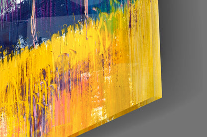 Close-up angled view of glass wall art showing yellow, orange, and deep blue brushstroke patterns with dripping paint effects.
