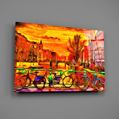 Colorful glass wall art depicting vibrant bicycles lined up against a fiery orange and yellow sunset cityscape with red buildings.