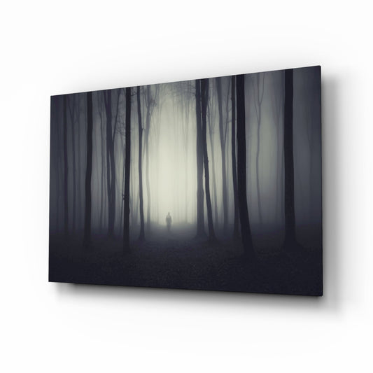 Angled view of glass wall art depicting a solitary figure standing in a misty forest corridor with dark trees and pale light, mounted against white background.