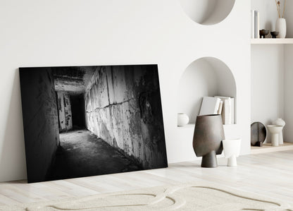 Black and white photograph of deteriorating concrete hallway with peeling walls displayed as glass wall art leaning against white wall.
