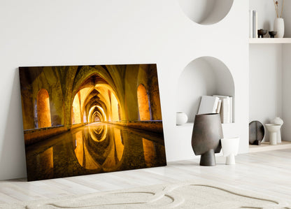 Glass wall art leaning against white wall displays golden-lit arched tunnel with water reflection in minimalist interior setting.