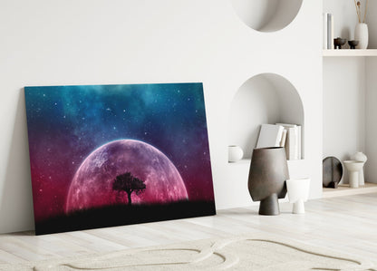 Glass wall art leaning against white wall displays silhouetted tree beneath glowing pink moon against starry blue gradient sky.