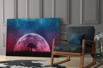 Mystical Night Glass Wall Art displayed in a modern living room beside a wooden chair, showing a pink glowing moon with silhouetted tree against a starry blue-to-purple gradient sky.