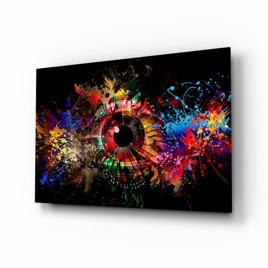 Angled perspective view of a glass wall art panel featuring a vibrant multicolored eye design with paint splatters radiating outward on black background.