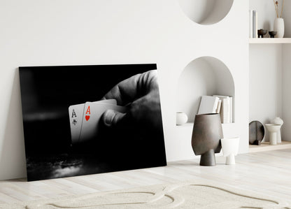 Black and white glass wall art depicting a hand holding two playing cards (Ace of Spades and Ace of Hearts) displayed in minimalist interior setting.