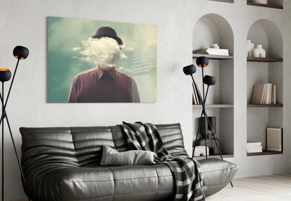 Surreal glass wall art depicting a man with bowler hat and cloud replacing his head, displayed above black leather sofa in modern living room setting.