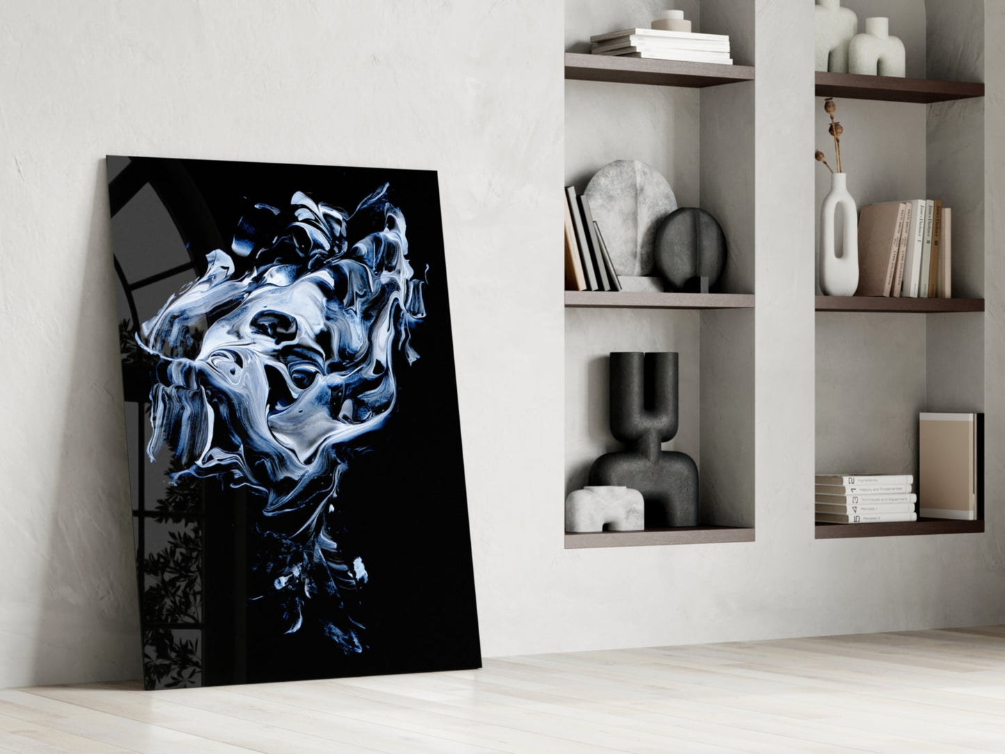 Abstract gray glass wall art leaning against white wall beside shelving unit displaying books and decorative objects.