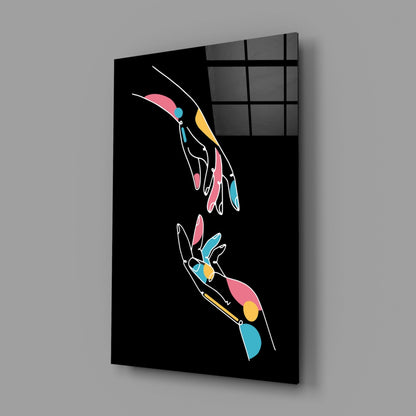 Black glass panel with two outlined hands in white holding colorful geometric shapes in pink, blue, and yellow, displayed at an angle against gray wall.