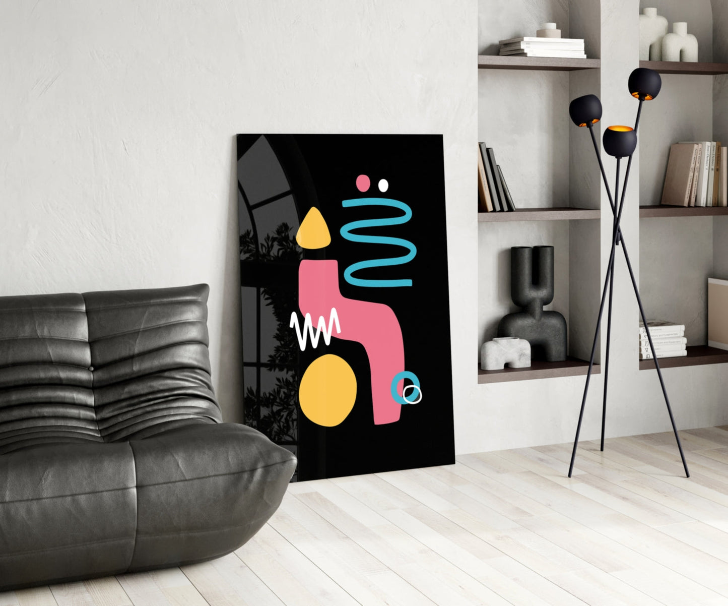 Modern living room interior featuring a black canvas with abstract geometric shapes in pink, yellow, and cyan, leaning against white wall beside gray leather chair.