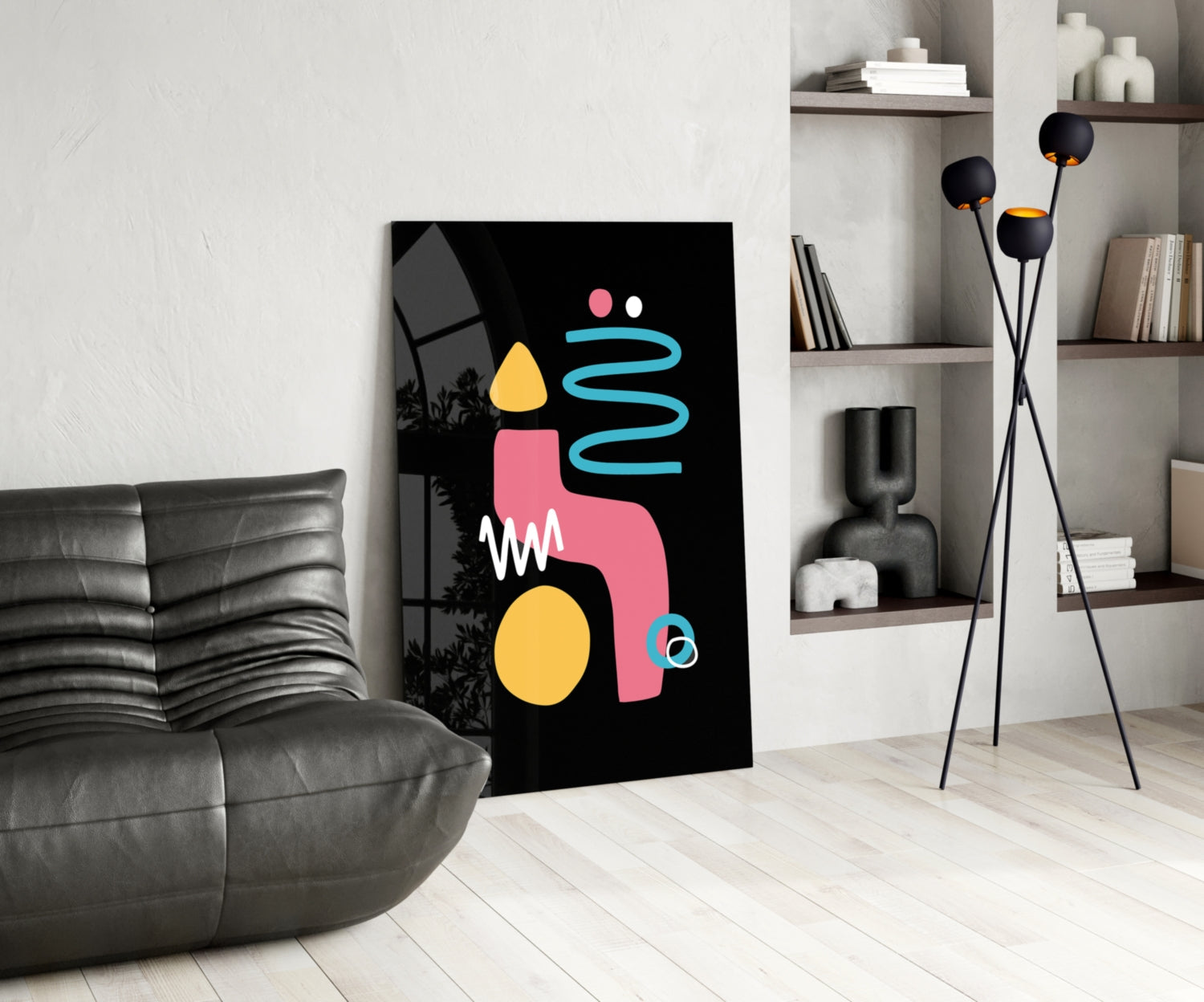 Modern living room interior featuring a black canvas with abstract geometric shapes in pink, yellow, and cyan, leaning against white wall beside gray leather chair.