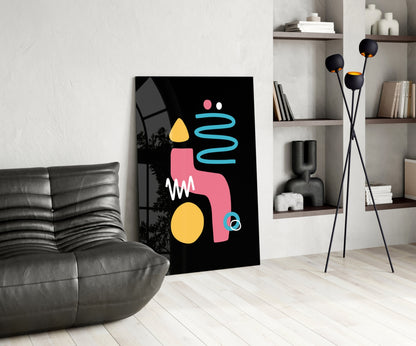 Modern living room interior featuring a black canvas with abstract geometric shapes in pink, yellow, and cyan, leaning against white wall beside gray leather chair.
