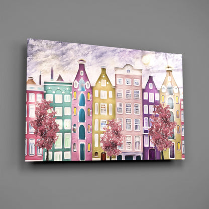 Colorful glass wall art featuring a row of stylized European buildings in pink, green, yellow, and purple with flowering trees against a cloudy sky.